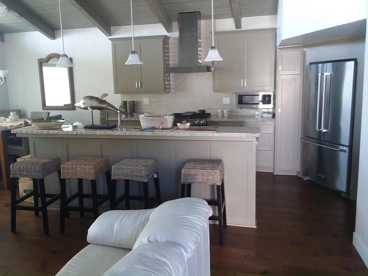A finished open-plan kitchen in pale greige shaker cabinets. A long
                  island with a speckled stone top and four woven bar stools faces a
                  run of counter with a stainless chimney hood over the range, a
                  herringbone tile backsplash behind it, and a tall stainless
                  refrigerator to the right. Painted ceiling beams run overhead and
                  the floor is dark hardwood.