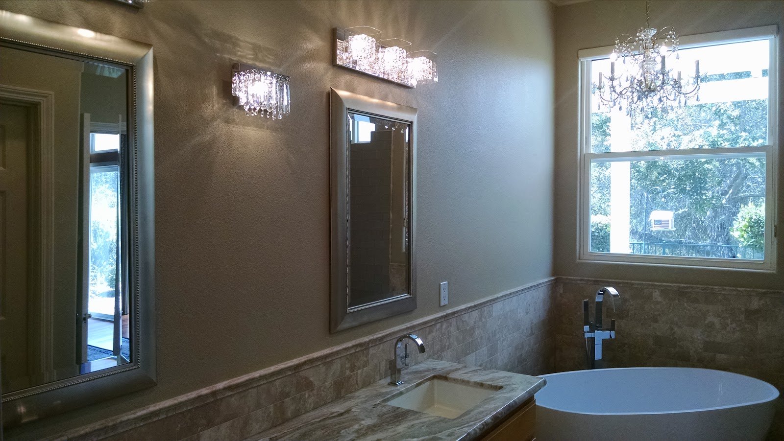 A finished master bathroom. A long marble vanity top with an undermount
            basin runs along a wall of travertine wainscot, under a framed mirror and
            a crystal bath bar. A second crystal sconce lights the near wall, a
            freestanding oval tub stands under a tall window at the far end, and a
            chandelier hangs above it with oak trees visible outside.
