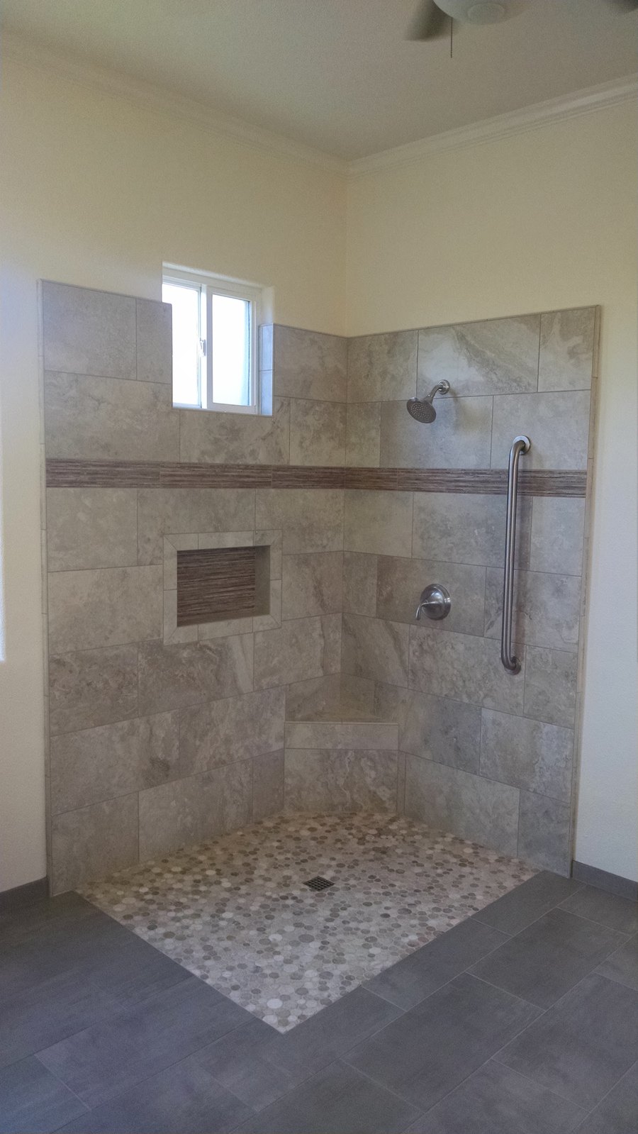 A finished curbless shower. Large gray-veined stone tiles line two
                  walls, broken by a narrow horizontal band of dark linear mosaic that
                  continues into a recessed soap niche. A stainless grab bar runs down
                  the right wall beside the valve, a tiled corner bench sits at the
                  back, and the floor is round pebble mosaic draining to a center
                  grate, level with the gray floor tile outside it.